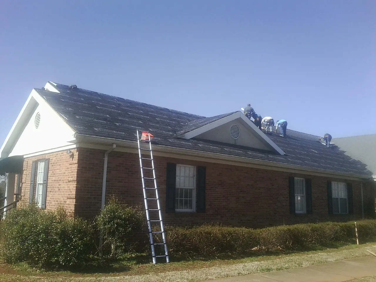 Crew tearing shingles off a brick ranch roof with a ladder set — Metal Roof Repair work by our crew in Cedarhurst