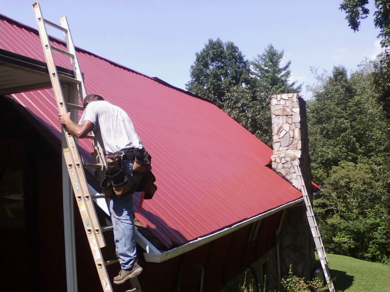 Roofer on a ladder beside a red metal roof and stone chimney — Soffit & Fascia Repair by Roofing Brothers in Cedarhurst